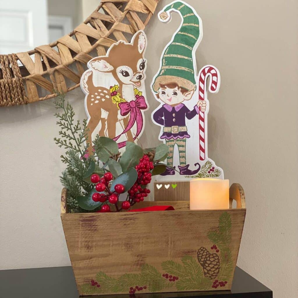 A finished set of an elf and reindeer shelf sitters behind a gold box holding a candle and Christmas greens for the blog post "Christmas Shelf Decor".