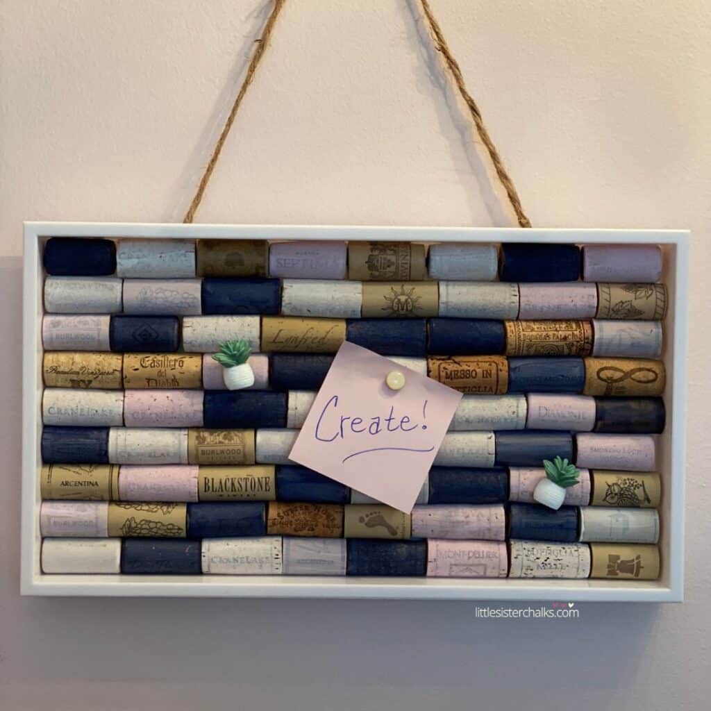 A navy/white/pink cork board hanging on a wall with a pink post-it note that says create and two small tacks that look like little plants in white post for the blog post "Create a Chic Wine Cork Board in 5 Easy Steps".