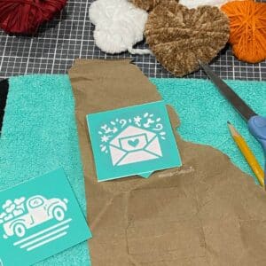 Craft supplies including mesh stencils, brown paper, a pencil, scissors and a teal microfiber cloth for the blog post "5 Easy Steps to Create Recycled Cardboard Hearts".