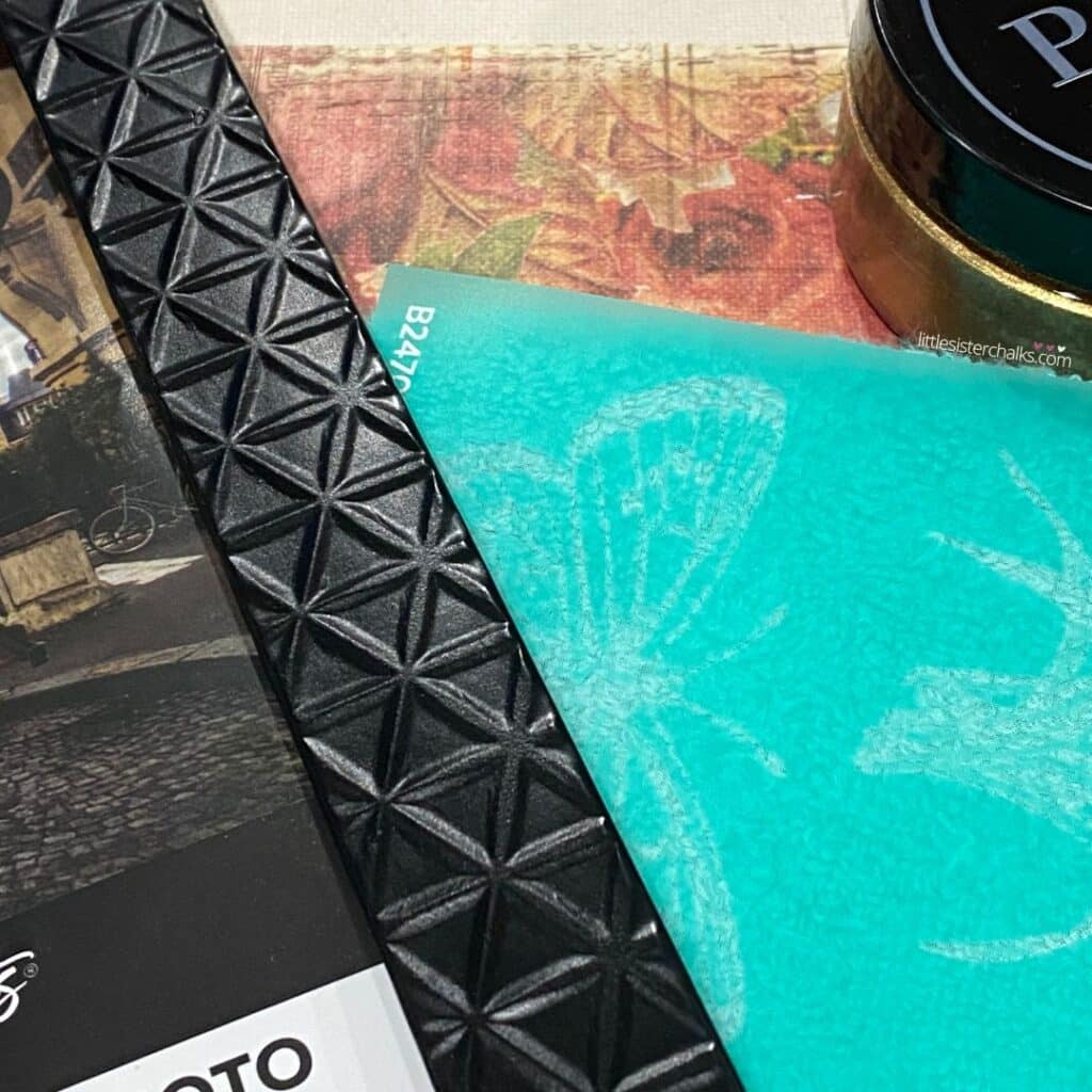 A close-up of a dark picture frame with a pattern, a stencil, floral scrapbook paper and a jar of gold chalk paste for the blog post "Transform Old Picture Frames into Handy Dry Erase Memo Boards".