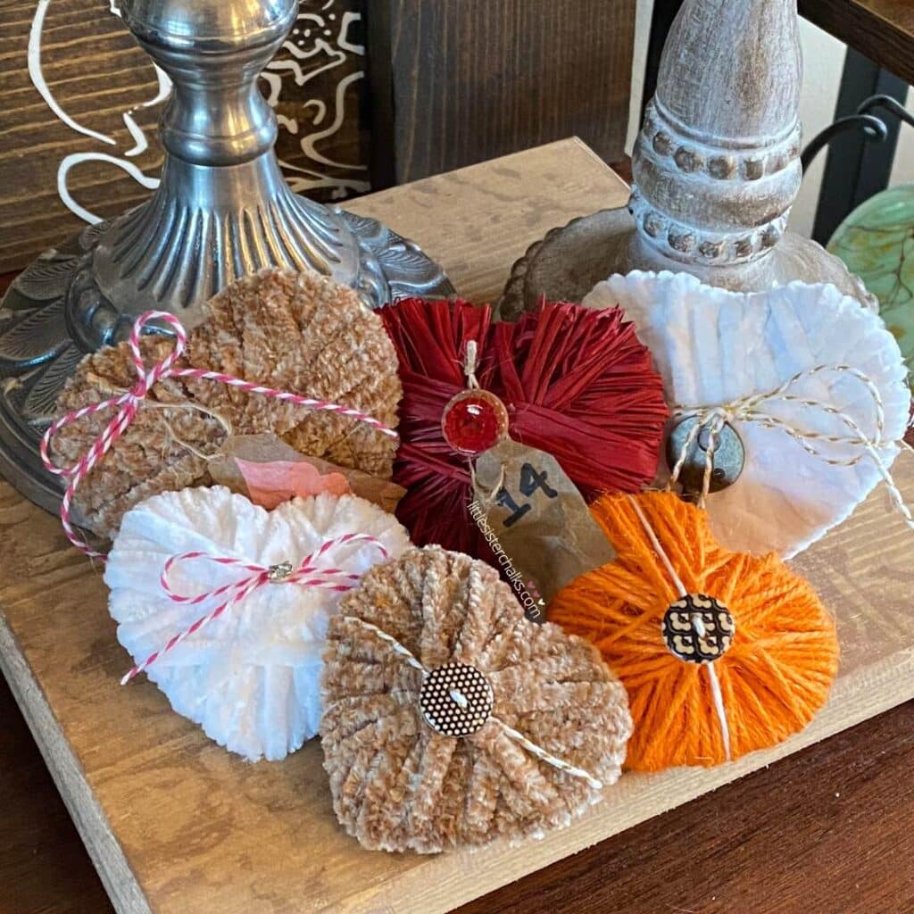 6 dimensional hearts made with yarn and twine in tan, white, orange and red on a wood riser with 2 candlesticks for the blog post "5 Easy Steps to Create Recycled Cardboard Hearts".