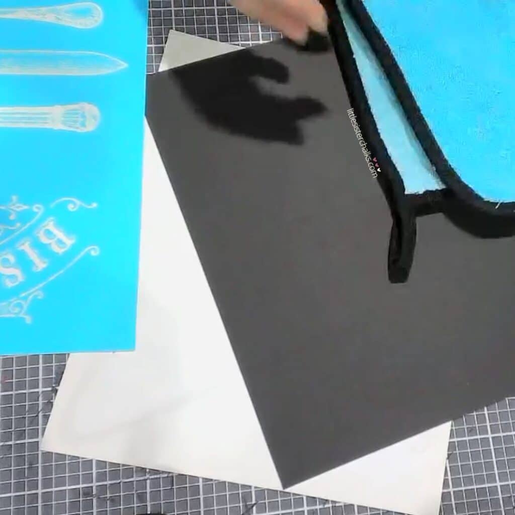 A close-up of a bistro cafe stencil, a piece of black cardstock, a white piece of paper and a teal microfiber cloth on a craft mat for the blog post "Transform Old Picture Frames into Handy Dry Erase Memo Boards".