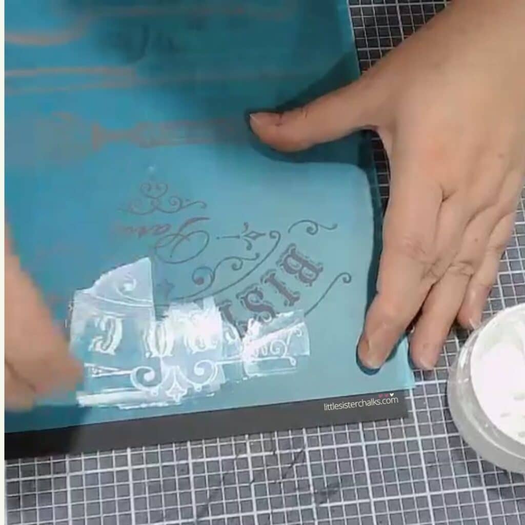 Two hands chalking a bistro cafe stencil in white chalk paste on a craft mat for the blog post "Transform Old Picture Frames into Handy Dry Erase Memo Boards".