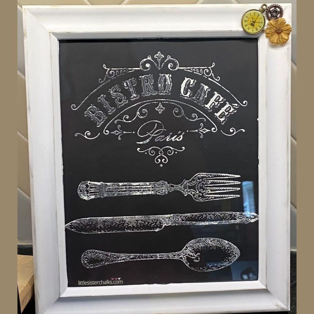 A white frame with a black insert with a white stenciled design that reads "Bistro Cafe Paris" with a fork, knife and spoon stacked over each other for the blog post "Transform Old Picture Frames into Handy Dry Erase Memo Boards".