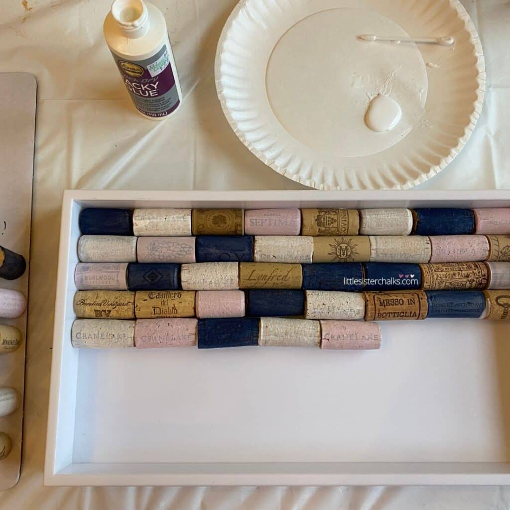 Natural and painted wine corks in navy, pink and white, being glued into a white tray with a bottle of tacky glue, a paper plate with glue and a swab on it for the blog post "Create a Chic Wine Cork Board in 5 Easy Steps".