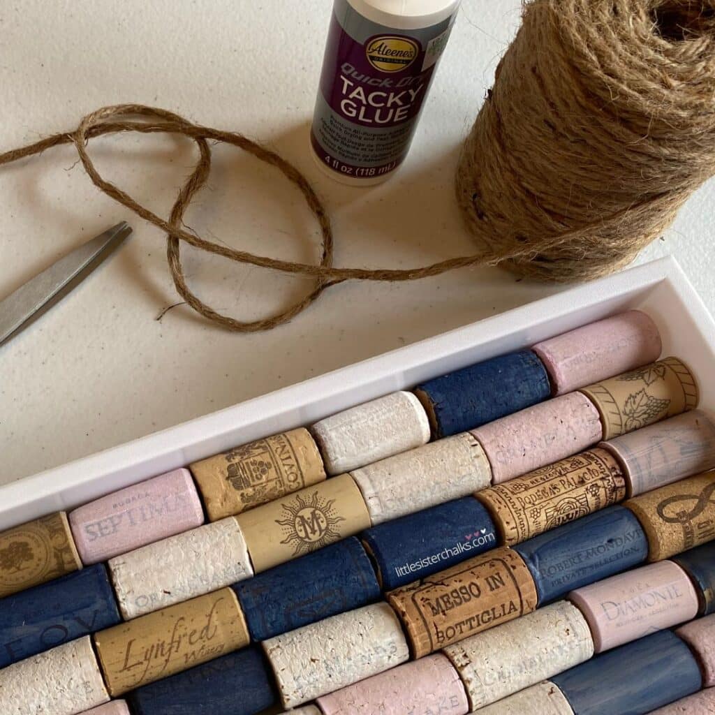 A close up of a tray holding natural and painted wine corks in navy, pink and white, with twine, scissors and glue above it for the blog post "Create a Chic Wine Cork Board in 5 Easy Steps".