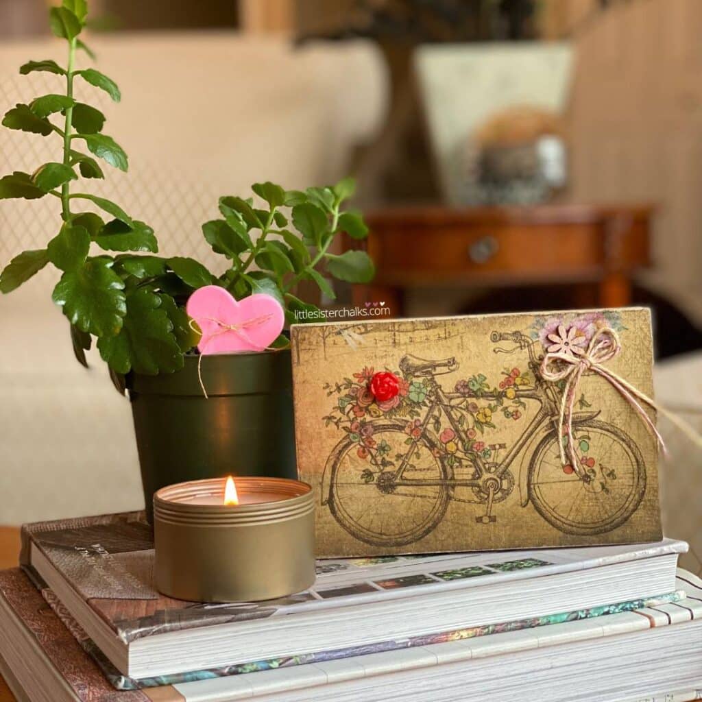 A vignette including a plant in a green pot with a pink foam heart staked in it, a gold candle and a bicycle decoupaged on a wood block and embellished with a red flower button, a wood flower and a small bow for the blog post "Valentine's Day Mixed Media Art Project".