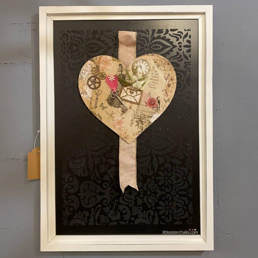 A framed chalkboard with a damask stenciled pattern as the background to a decoupaged and embellished heart and pink and gold ribbon for the blog post "Valentine's Day Mixed Media Art Project".