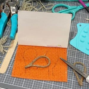 A photo of an orange piece of fabric, two tear drop shapes made from jute, and scissors on a craft mat for the blog post "Crafting Beyond the Bin: Adorable Bunny in a Bucket Upcycle from a Laughing Cow Cheese Container".