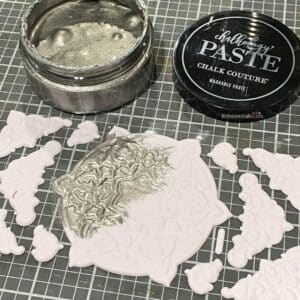 A photo of Chalk Couture Artisan Add-ons on a craft mat being painted in silver Chalkology Paste for the blog post "Crafting Beyond the Bin: DIY Faux Rusted Tin Cans".