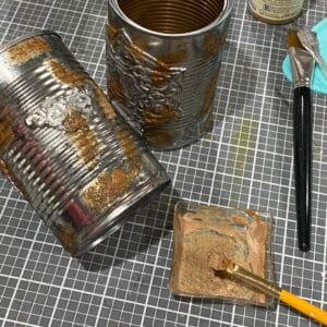 Two tin cans on a craft mat with a glue and cinnamon faux rust mixture being applied with a yellow paint brush for the blog post "Crafting Beyond the Bin: DIY Faux Rusted Tin Cans".