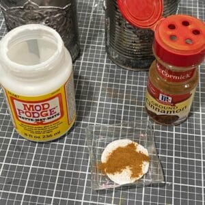 A bottle of Mod Podge and a container of ground cinnamon with some of each in a color tray for the blog post "Crafting Beyond the Bin: DIY Faux Rusted Tin Cans".