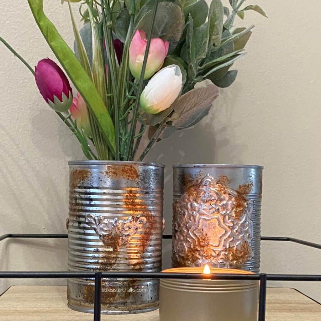 Two embellished tin cans, one holding fake greens and purple and white tulips, with a burning candle in front of them in a wood and metal tray for the blog post "Crafting Beyond the Bin: DIY Faux Rusted Tin Cans".