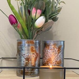 Two embellished tin cans, one holding fake greens and purple and white tulips, with a burning candle in front of them in a wood and metal tray for the blog post "Crafting Beyond the Bin: DIY Faux Rusted Tin Cans".