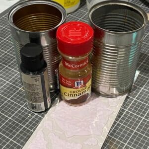 A photo of 2 cans, black acrylic paint, cinnamon, and a package of Chalk Couture artisan add-ons on a craft mat for the blog post "Crafting Beyond the Bin: DIY Faux Rusted Tin Cans".