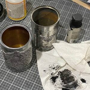 A photo of 2 embellished tin cans being "aged" with black acrylic paint and a piece of a plastic grocery bag for the blog post "Crafting Beyond the Bin: DIY Faux Rusted Tin Cans".