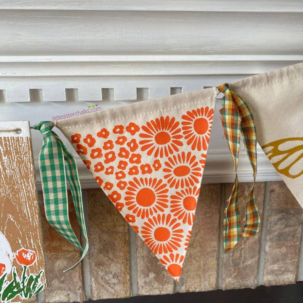 A photo of a piece of a spring garland hanging from a white mantel featuring the Full Bloomin' stencil from the Chalk Couture x Tiffani Thiessen Collaboration chalked in orange.