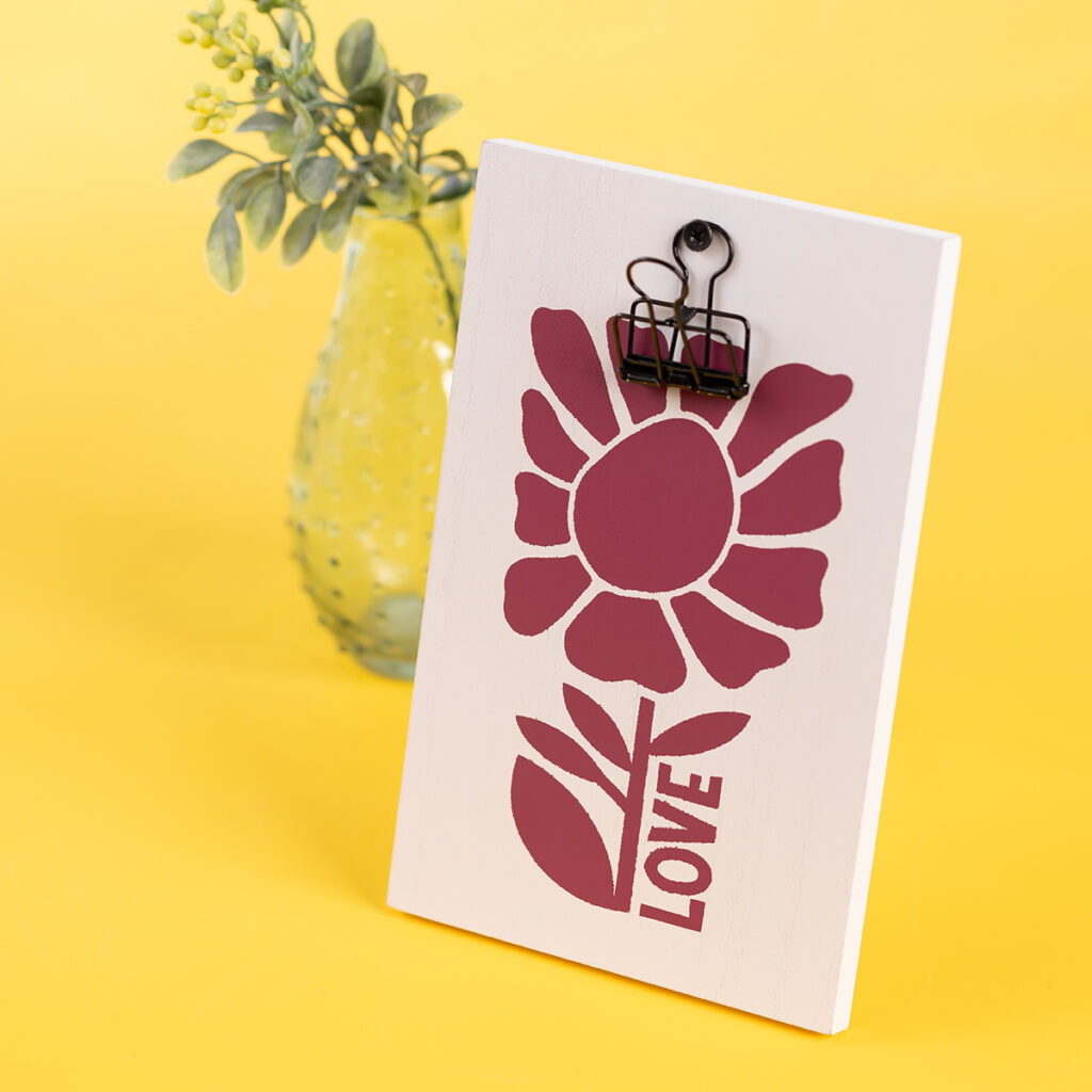 The finished project from the Chalk Couture Love Blooms Try Me Kit which is a white clipboard with the word "love" and a flower chalked in Currant Jam paste flanked by a vase with small flowers for the blog post "Try your hand at chalking with a Chalk Couture Try-me Kit".