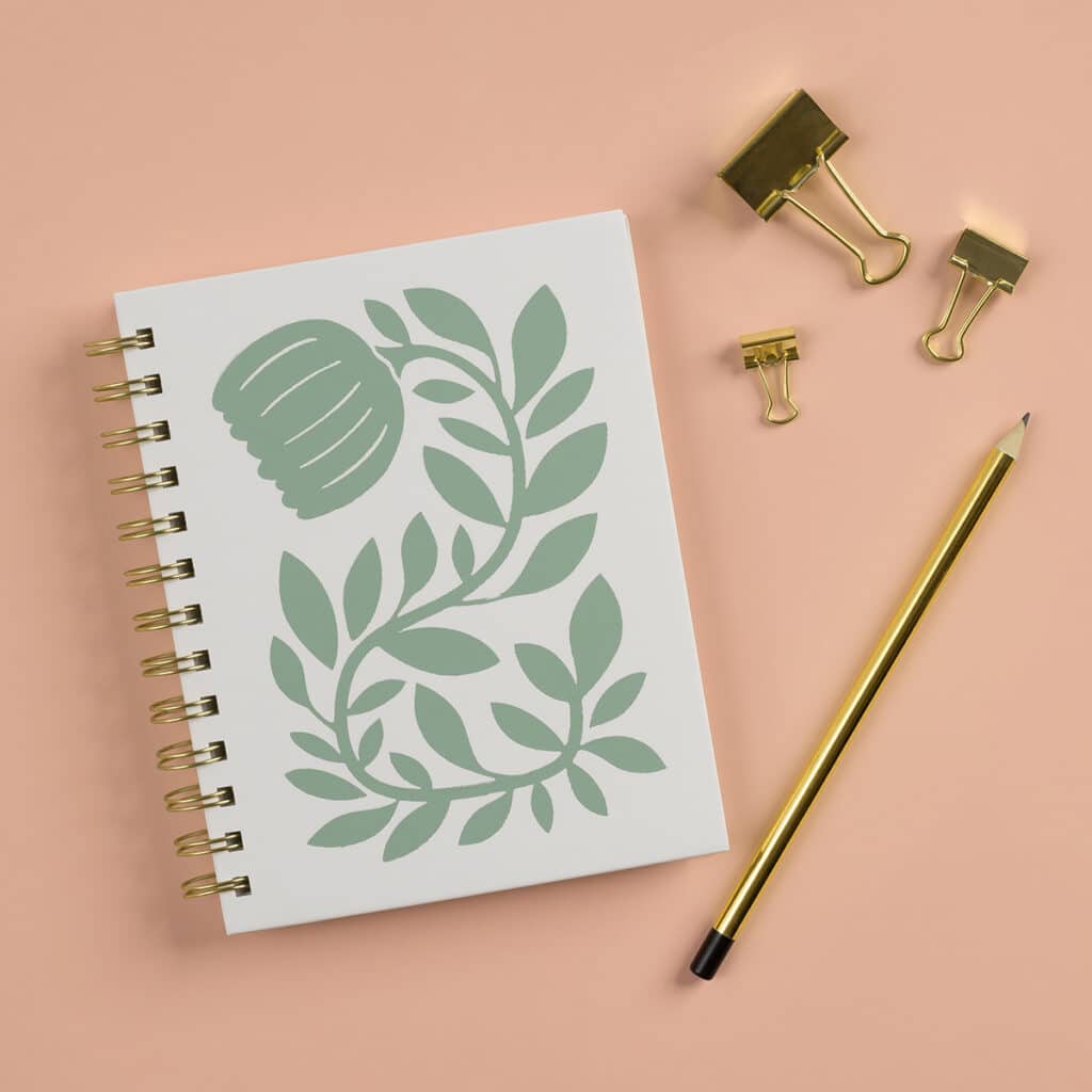 A photo of the finished Chalk Couture Wandering Vine Try Me Kit featuring a white notebook with the Wandering Vine chalked in Eucalyptus on the cover flanked by a gold pencil and gold binder clips for the blog post "Try your hand at chalking with a Chalk Couture Try-me Kit".