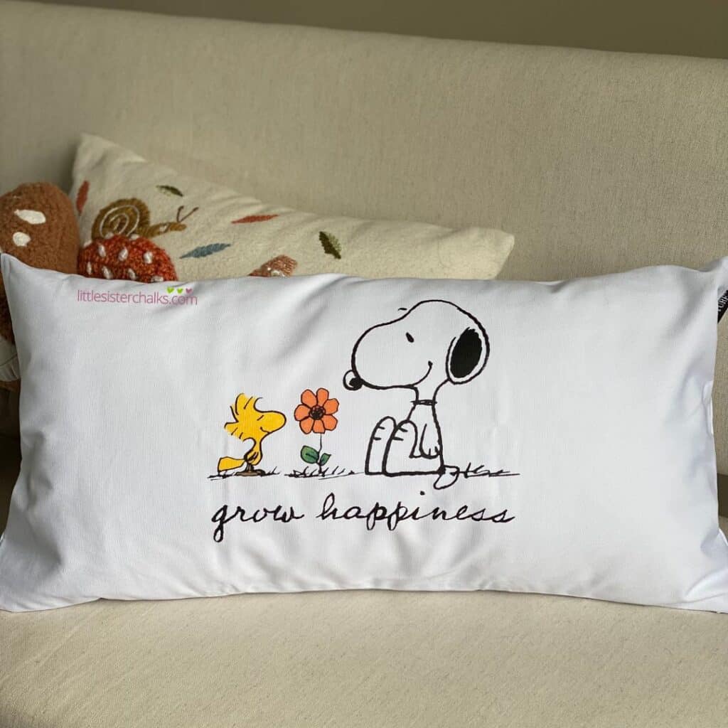 A white lumbar pillow with the Chalk Couture x Peanuts - Grow Happiness design inked on the pillow in Black Velvet ink for the blog post "Outdoor Fabric Accessories".