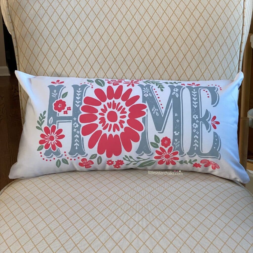 A white lumbar pillow with the word "Home" surrounded by flowers and leaves inked on the fabric in Ocean Mist and Couture Coral Chalk Couture Ink for the blog post "Outdoor Fabric Accessories".