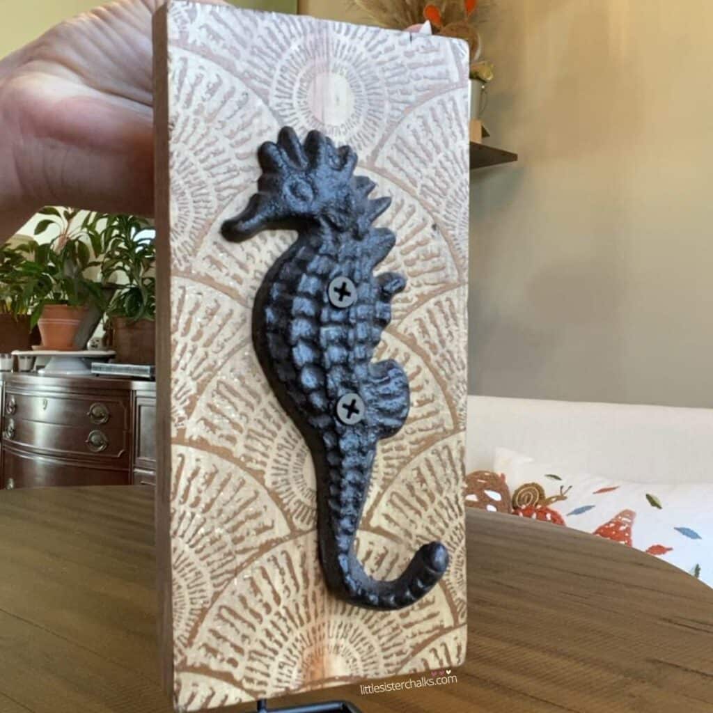 A photo of a piece of scrap wood decorated with a chalked pattern that looks like sea shells and an iron sea horse hook attached to it with screws for the blog post "Decorating with Metals".