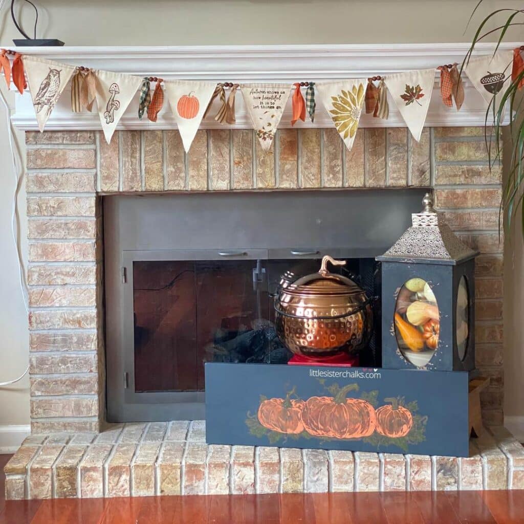 A photo of a hammered copper pumpkin flanked by a lantern with a silver top and a black wood sign with three pumpkins in front of a fireplace with a fabric banner hanging from the mantel for the blog post "Decorating with Metals".